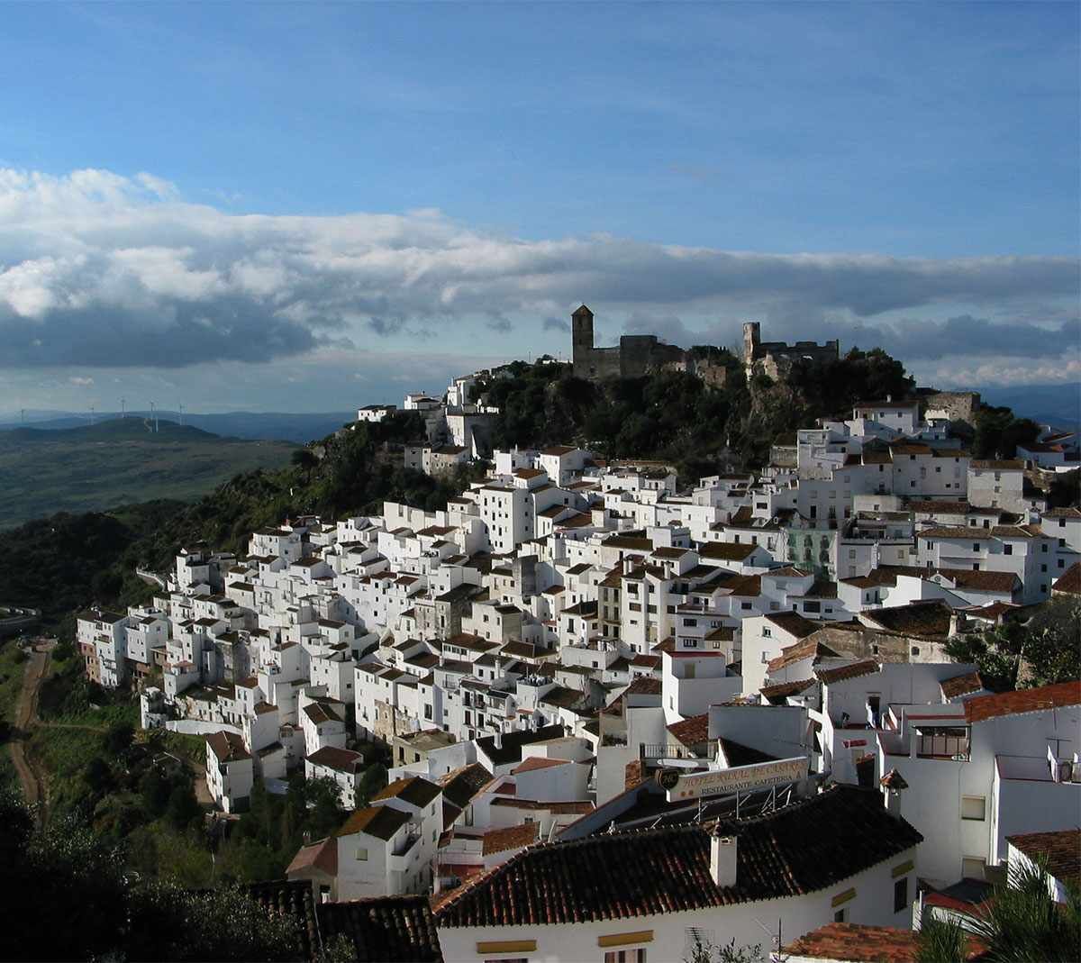 A densely packed white building area with a prominent tower on a hill, surrounded by greenery and a clear sky.