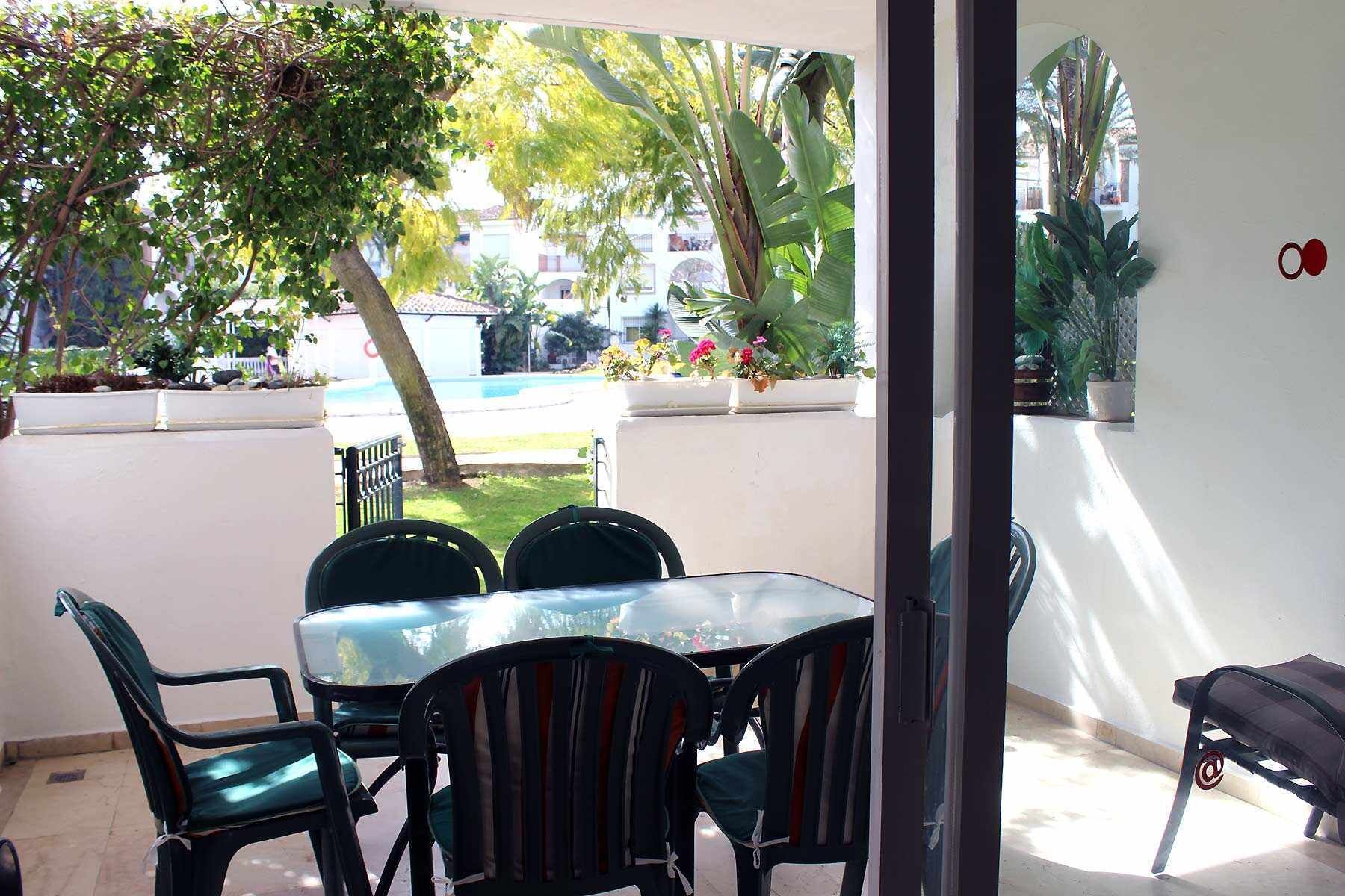Un patio con mesa y sillas, rodeado de plantas en macetas y vista a una piscina.