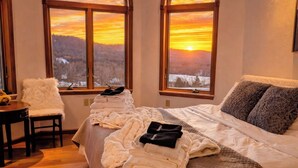 Een hotelkamer met een bed, een stoel en uitzicht op een ondergaande zon boven een bebost landschap.