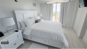 Un dormitorio blanco con cama, mesita de noche y vista al mar.