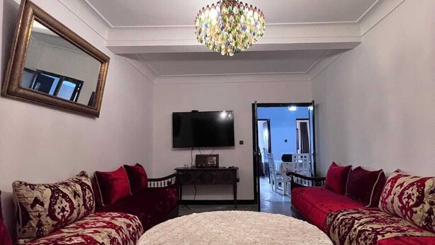 Un salón con dos sofás rojos, una mesa de centro redonda, una araña, un televisor de pantalla plana y un espejo en la pared.