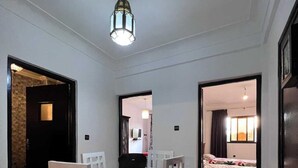 Comedor con mesa y sillas, una lámpara en el techo y vista hacia un dormitorio con cama y cortinas.