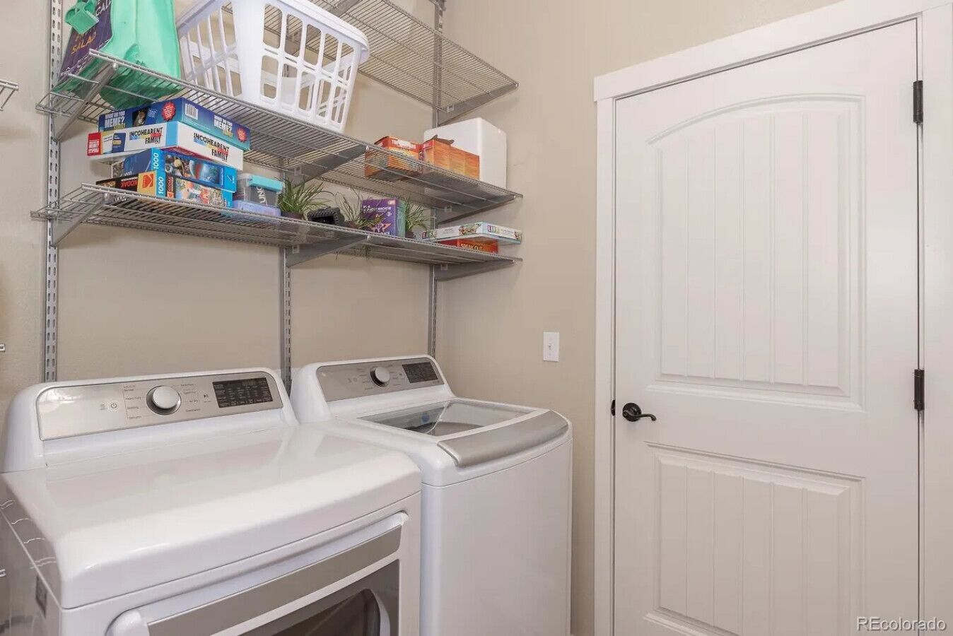 A laundry room with two white front-loading washers and dryers, a white door, and shelves above holding various items including a basket and boxes.