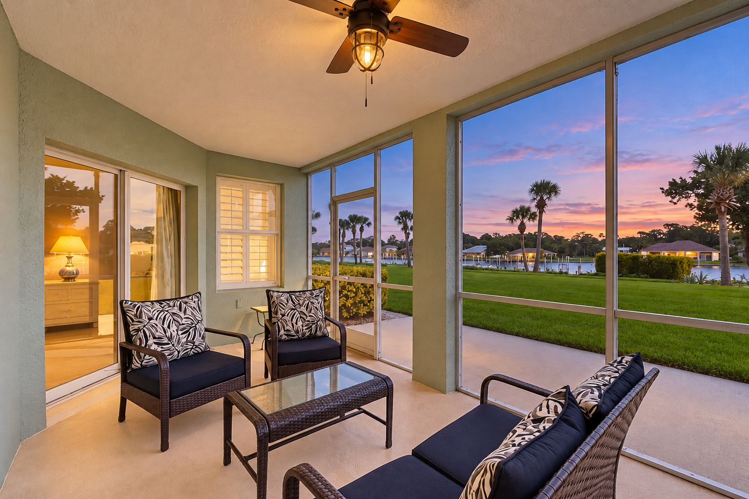 A sunlit room with a glass table, two chairs, and a sofa, overlooking a golf course at sunset.