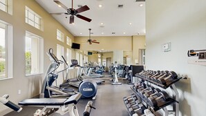 Academia com diversos equipamentos de exercício, ventiladores de teto e janelas amplas.