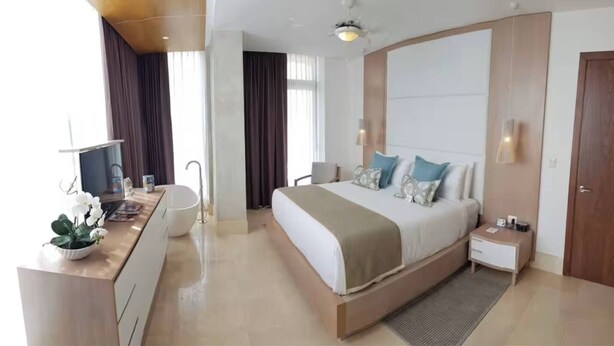 Quarto de hotel moderno com uma cama grande, mesas de cabeceira, uma escrivaninha com cadeira, uma janela ampla e um ventilador de teto.