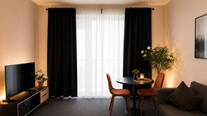 Una habitación de hotel moderna con televisor de pantalla plana, un sofá, una mesa de comedor con sillas y un ventanal con cortinas translúcidas.