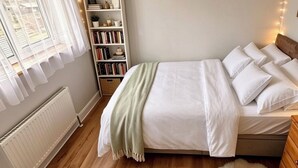 A bedroom with a bed, a bedside table, a chair, a radiator, a bookshelf, and string lights.