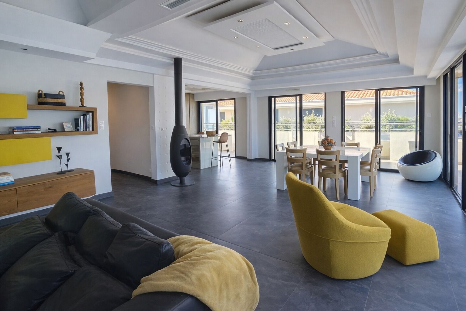 A spacious living area with a modern sofa, yellow ottoman, and a dining table with chairs. A unique black spherical chair is visible in the background.