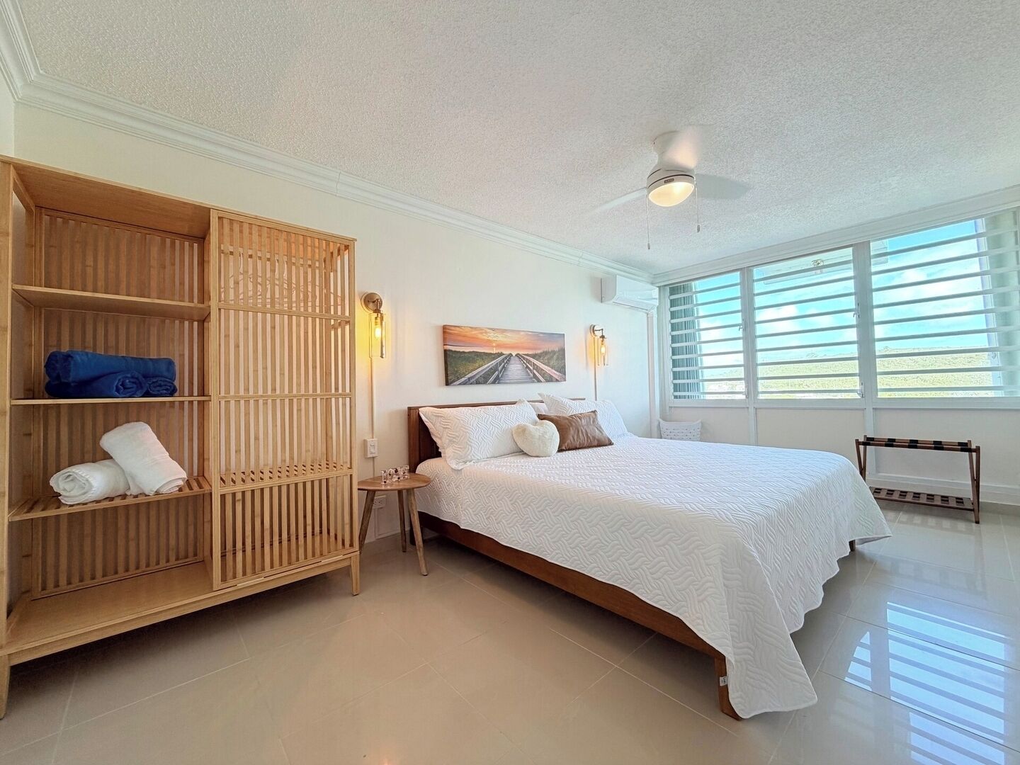 Une chambre à coucher comprenant un lit, une armoire en bois, une table de chevet, une lampe fixée au mur et une fenêtre avec des stores.