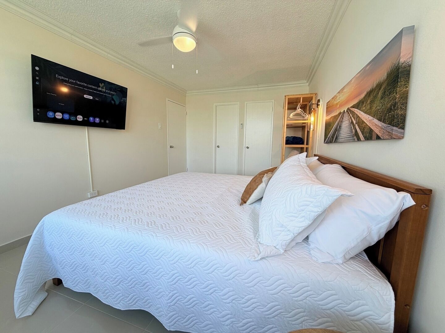 A neatly made bed with a white quilt, a television mounted on the wall, and a painting of a beach scene.