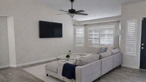 Una sala de estar con un sofá gris, una mesa de centro, un ventilador de techo y un televisor montado en la pared.