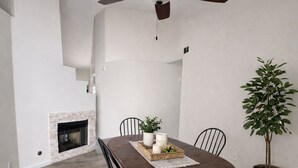 Un comedor con una mesa redonda de madera, sillas negras, un ventilador de techo, una chimenea y una planta en maceta.