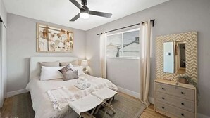 Un dormitorio con cama, mesitas de noche, ventilador de techo, ventana con persianas, cómoda y espejo.