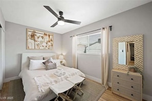 Ein Schlafzimmer mit Bett, Nachttischen, Deckenventilator, Fenster mit Jalousien, Kommode und Spiegel.