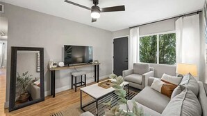 Una sala de estar moderna con una televisión de pantalla plana, un ventilador de techo, un sofá, una mesa de centro y un amplio ventanal con vistas a la vegetación.
