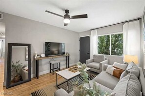 Ein modernes Wohnzimmer mit Flachbildfernseher, Deckenventilator, Sofa, Couchtisch und großem Fenster mit Blick auf Grünflächen.