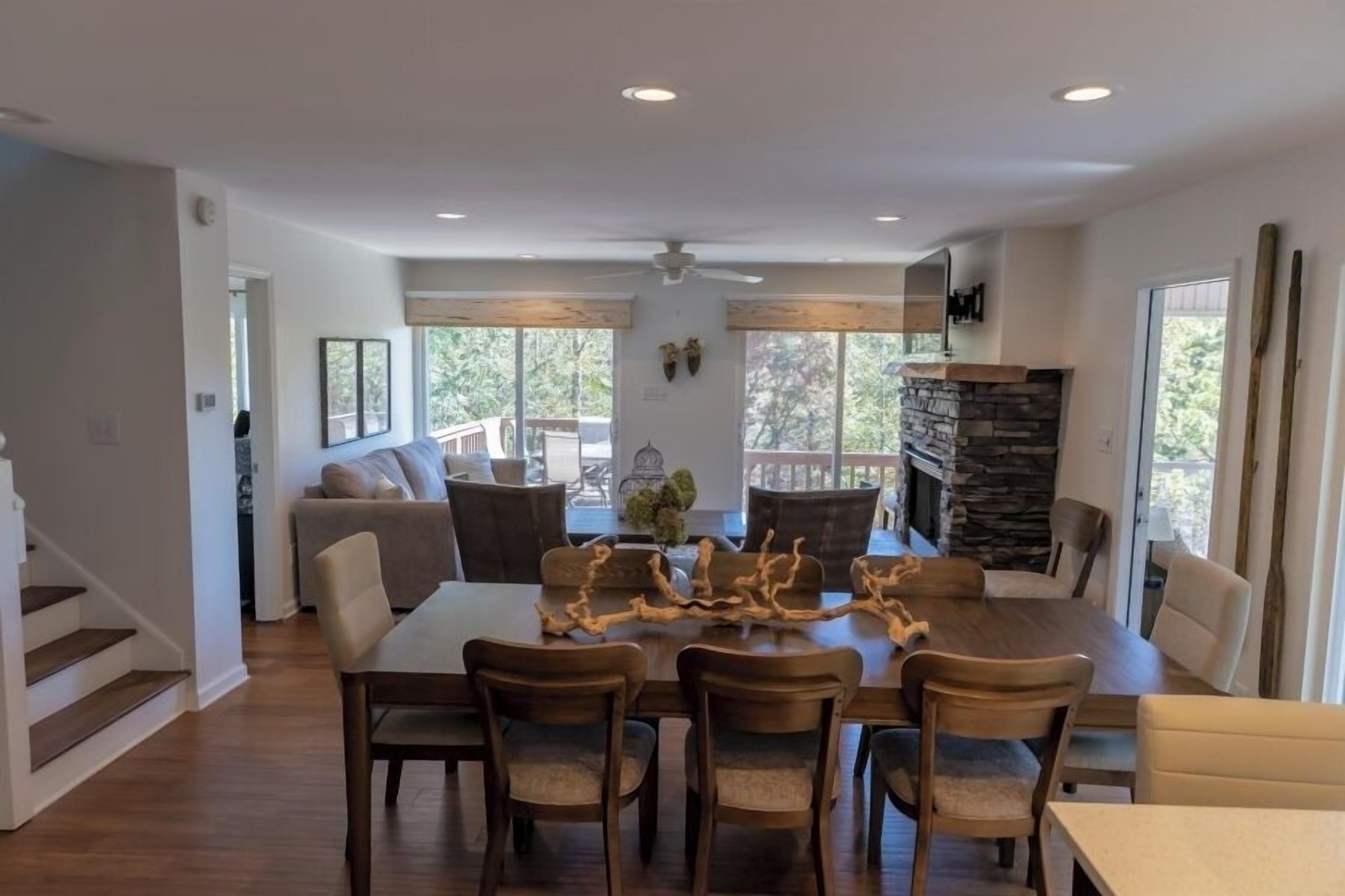 A spacious dining area with a large wooden table and chairs, a stone fireplace, and large windows offering a view of the outdoors.