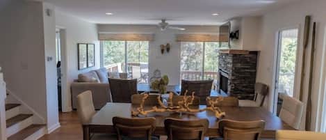 A spacious dining area with a large wooden table and chairs, a stone fireplace, and large windows offering a view of the outdoors.