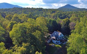 Uma casa de dois andares rodeada por uma floresta densa, com uma montanha ao fundo.
