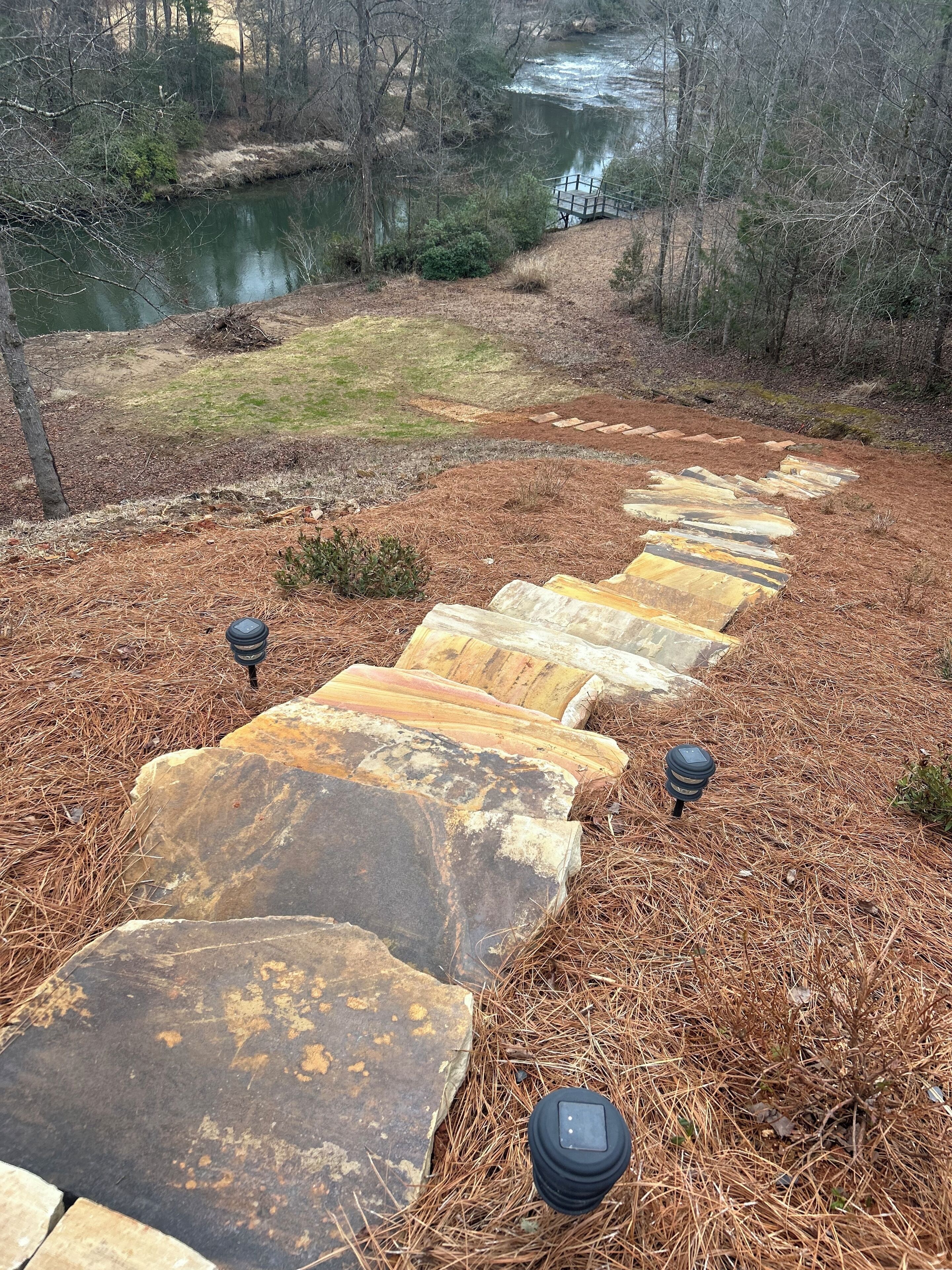 A stone pathway leading to a body of water, surrounded by pine straw and trees.