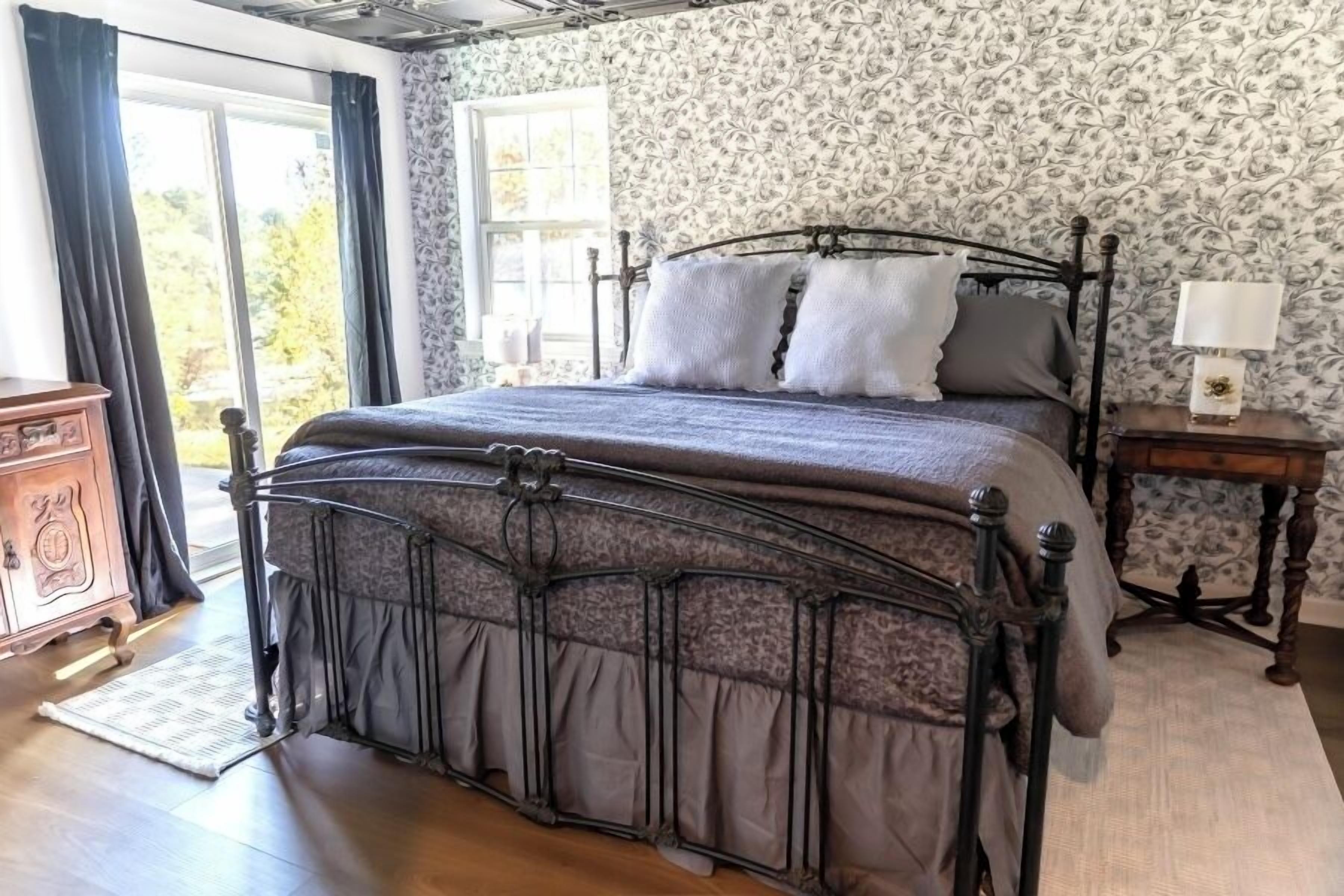 A bedroom with a large bed, a wooden dresser, a nightstand with a lamp, and a window with curtains.