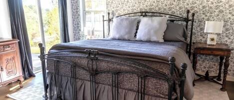 A bedroom with a large bed, a wooden dresser, a nightstand with a lamp, and a window with curtains.