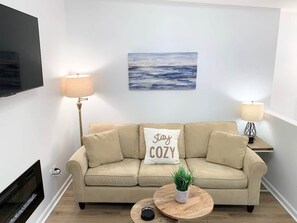 Un salon avec un canapé beige, une table basse ornée d’une plante, deux lampes, un téléviseur à écran plat et un tableau accroché au mur.