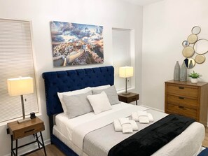 A bedroom with a large bed, two bedside tables, a headboard, a wall-mounted painting, a dresser, and a mirror.