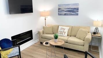 A living room with a flat-screen TV, a beige sofa with a 'stay cozy' pillow, a small round coffee table, and a lamp.