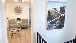 Una sala de estar con un reloj de pared redondo, dos sillones estampados, una mesita y un cuadro de un paisaje urbano.