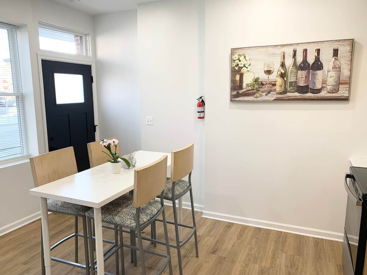 A dining area with a white table and four chairs, a black door, and a fire extinguisher mounted on the wall.