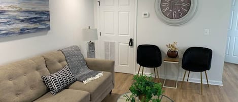 A living room with a beige sofa, two black chairs, a glass coffee table with a plant, a wall clock, and a door with a white frame.