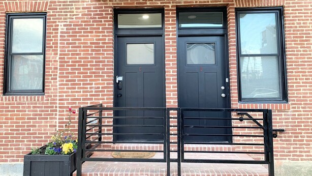 A two-story brick building with a black front door, two windows on the upper floor, and a small porch with a black railing.