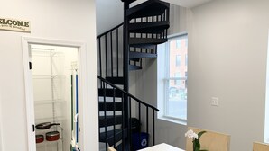 A modern apartment with a white interior, a black spiral staircase, a dining area with a white table and chairs, and a view of the outside through a window.