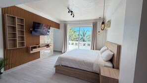 A bedroom with a large bed, a TV mounted on a wooden panel wall, a balcony with a view of trees, and a ceiling light fixture. - Sea View Room - Superior Panoramic No. 4 (Playa Juan Dolio)