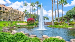 A landscaped garden with a central water feature, palm trees, and a building with balconies. - Beach, Pool & Pickleball Short Walk to Beach! (Kihei)