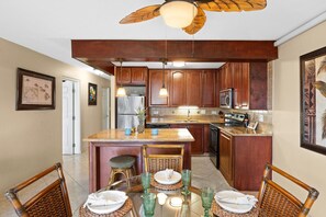 A kitchen with wooden cabinets, a central island, and a dining area with wicker chairs and a glass table. - Beach, Pool & Pickleball Short Walk to Beach! (Kihei)