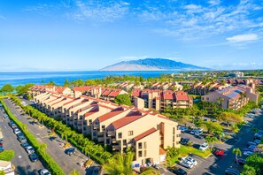 A resort with multiple buildings, a parking lot, and a mountain in the background. - Beach, Pool & Pickleball | Maui Kihei Condo (Kihei)