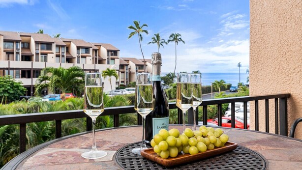 A table with a bottle of champagne, glasses, and a bowl of grapes on a balcony overlooking a resort with palm trees and a view of the ocean. - Beach, Pool & Pickleball Short Walk to Beach! (Kihei)