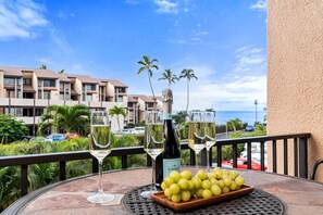 A table with a bottle of champagne, glasses, and a bowl of grapes on a balcony overlooking a resort with palm trees and a view of the ocean. - Beach, Pool & Pickleball Short Walk to Beach! (Kihei)