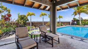 Un espacio al aire libre techado con piscina, sillas de descanso y vistas a palmeras y un paisaje rocoso.