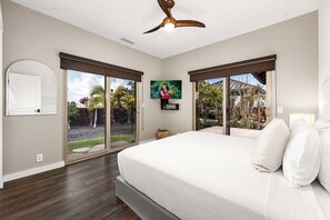 A bedroom with a large bed, a ceiling fan, a TV, and a sliding glass door leading to a garden. - 🏖️Private Luxury Home at Waikoloa Beach Resort | Pool, Outdoor TV, Sleeps 8 (Waikoloa)