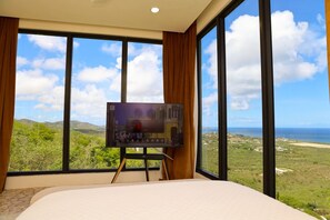 A room with a large bed, a television on a stand, and a scenic view through large windows. - Ocean-View 2BR + Soaking Pool · Christiansted (Christiansted)
