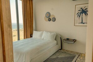 A bedroom with a large bed, a view of the outdoors, a small bedside table, and a wall decoration. - Ocean-View 2BR + Soaking Pool · Christiansted (Christiansted)