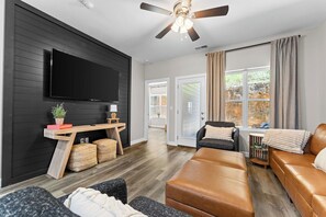 A modern living room with a flat-screen TV, a ceiling fan, and a large window with curtains. - New! 3 Bedroom Condo, Sleeps 8, No Stairs, Near Strip (Branson)