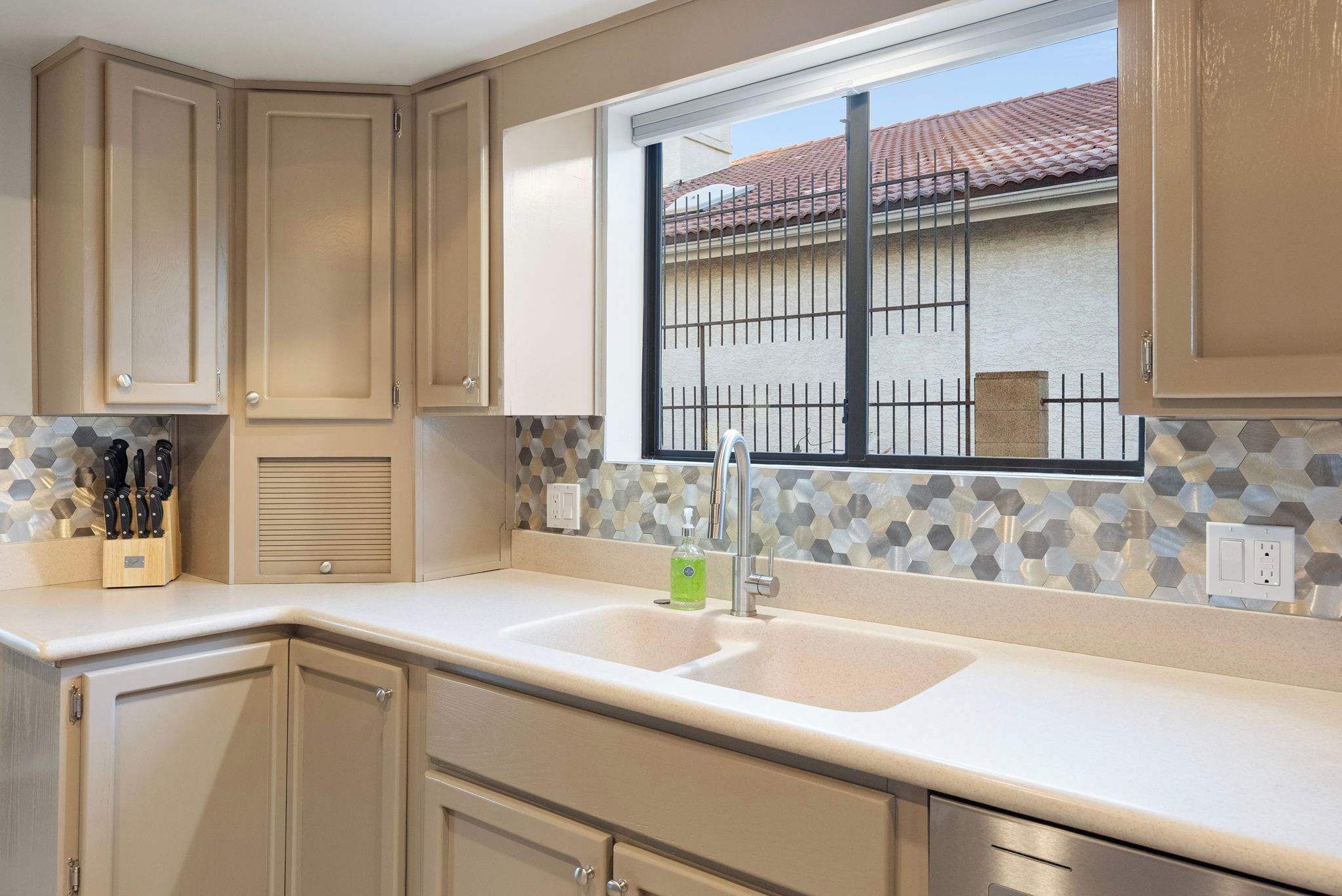 A kitchen with beige cabinets, a double sink, and a window with a view of a house and a fence.