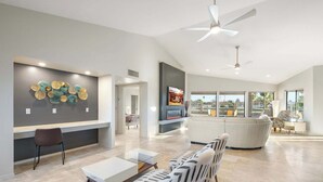 Ein geräumiges Wohnzimmer mit einem modernen Sofa, einem Couchtisch und einem an der Wand montierten Fernseher. Es gibt einen Deckenventilator, einen Kamin und ein großes Fenster mit Blick ins Freie.