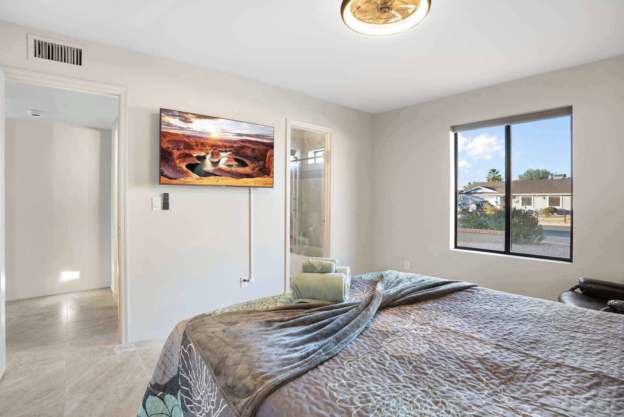 A bedroom with a large bed, a TV mounted on the wall, a window with a view of the outside, and a door leading to another room.