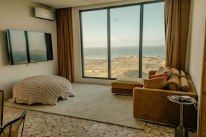 Ein Zimmer mit großem Fenster, das Meer und Hügel zeigt, eine Couch, ein kleiner Tisch mit Lampe sowie ein an der Wand befestigter Fernseher.